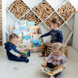 Pu&ograve; includere: Tre bambini sono seduti sul pavimento a leggere libri. I libri sono su una libreria di legno con il nome "Alexis" scritto su di essa. La libreria &egrave; piena di libri per bambini colorati. I bambini indossano abiti casual. Lo sfondo &egrave; una parete bianca con una parete di legno dietro.