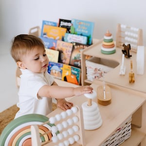 May include: A child plays with a wooden toy stacking ring set, a rainbow stacking toy, and a wooden abacus. The toys are on a wooden table with a bookshelf in the background.
