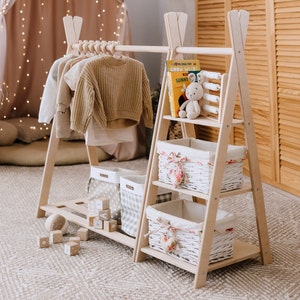 Puede incluir: Un perchero de madera en forma de A con un estante superior de madera blanca y dos estantes inferiores. Los estantes están llenos de cestas de mimbre blancas con detalles florales. El perchero tiene una barra de madera para colgar la ropa. Hay varias prendas colgadas en la barra, incluyendo un suéter marrón y una camisa blanca. También hay varios bloques de madera en el suelo frente al perchero.