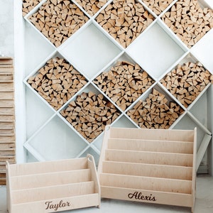 Pu&ograve; includere: Due librerie in legno con i nomi "Taylor" e "Alexis" incisi su di esse. Le librerie sono in legno chiaro e hanno un design semplice. Sono posizionate su un pavimento di piastrelle bianche di fronte a una parete bianca con un motivo a rombi riempito di legna da ardere impilata.