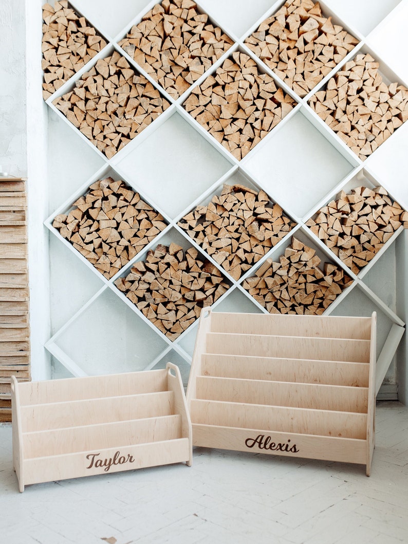 Pu&ograve; includere: Due librerie in legno con i nomi "Taylor" e "Alexis" incisi su di esse. Le librerie sono in legno chiaro e hanno un design semplice. Sono posizionate su un pavimento di piastrelle bianche di fronte a una parete bianca con un motivo a rombi riempito di legna da ardere impilata.