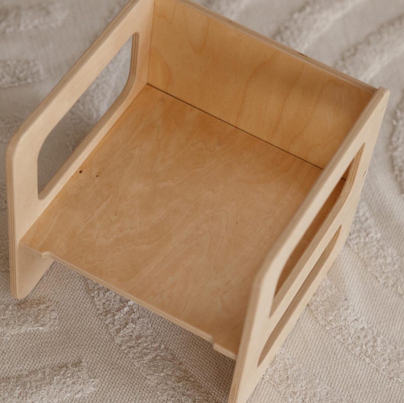 May include: A natural wood-toned step stool with a rectangular design. The stool features a flat top surface and two side handles for easy carrying. The wood grain is visible, and the stool is set on a textured, off-white surface.