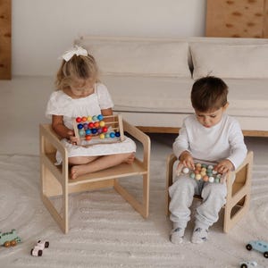 May include: Two children seated on small wooden chairs, each holding an abacus. One abacus has colorful beads, the other has neutral-toned beads. The chairs are light-colored wood, and the background includes a sofa and a patterned rug.