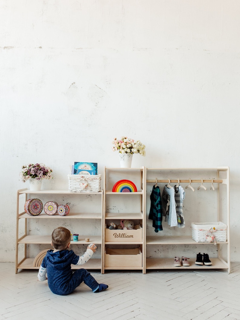 May include: A light wood children's clothing rack and shelf unit with a rainbow, toys, and books. The unit has a shelf labeled "William". A child is sitting on the floor in front of the unit.