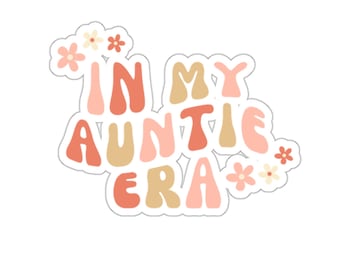 May include: A die-cut sticker with the phrase "IN MY AUNTIE ERA" in retro-style lettering. The text is in shades of peach, tan, and coral, with small floral accents. The sticker has a white border.