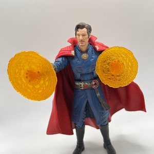 May include: A red-robed action figure of Doctor Strange, a Marvel superhero, holding two glowing orange magic discs. The figure has a blue and black costume with a red cape.
