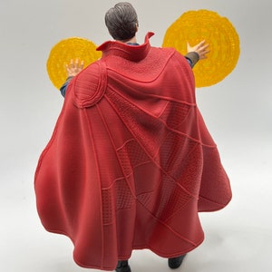 May include: A red and black action figure of Doctor Strange from the Marvel Cinematic Universe. The figure is wearing a red cape and holding two golden discs in his hands.