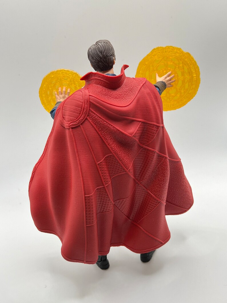 May include: A red and black action figure of Doctor Strange from the Marvel Cinematic Universe. The figure is wearing a red cape and holding two golden discs in his hands.