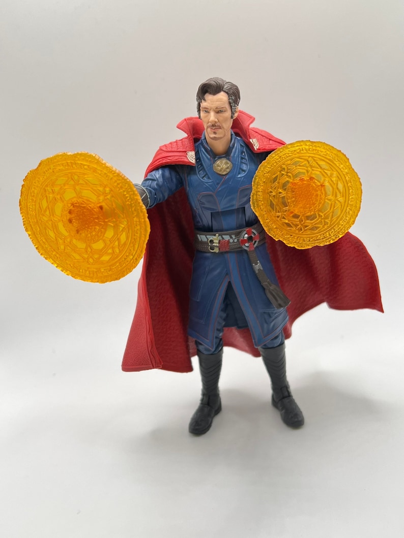 May include: A red-robed action figure of Doctor Strange, a Marvel superhero, holding two glowing orange magic discs. The figure has a blue and black costume with a red cape.