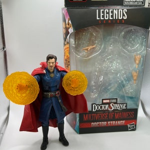 May include: Doctor Strange action figure from the Marvel Legends Series, holding two golden magic rings. The figure is wearing a red cape, blue robe, and black boots. The packaging features the Marvel Studios logo and the text "Doctor Strange in the Multiverse of Madness".