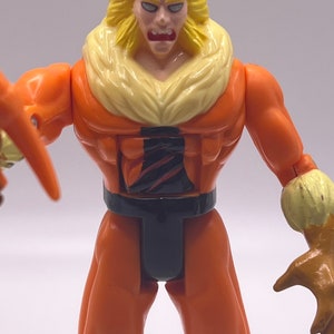 Puede incluir: Una figura de acci&oacute;n de pl&aacute;stico del personaje de Marvel Comics Sabretooth. La figura es naranja y amarilla con detalles negros. Sabretooth tiene una expresi&oacute;n feroz con dientes y garras afiladas.