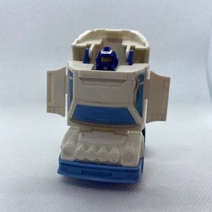 May include: A white and blue toy truck with a blue robot figure in the driver's seat. The truck has a blue windshield and white headlights. The robot has a blue helmet and a blue body.