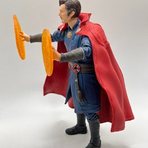 May include: A red-robed action figure of Doctor Strange, holding two glowing orange discs in his hands. The figure is posed in a dynamic stance, with one leg forward and the other back. The figure is wearing a blue and black costume with a red cape.
