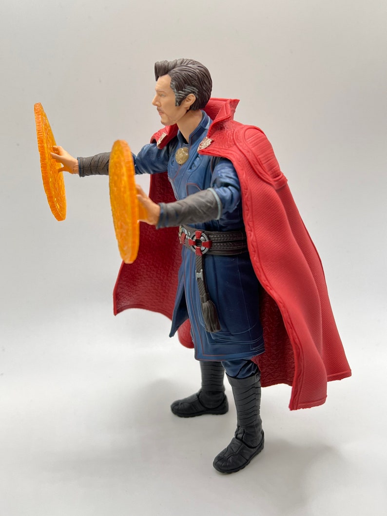May include: A red-robed action figure of Doctor Strange, holding two glowing orange discs in his hands. The figure is posed in a dynamic stance, with one leg forward and the other back. The figure is wearing a blue and black costume with a red cape.