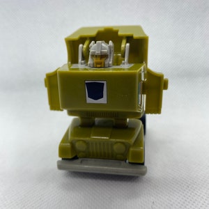 May include: A green and gray toy robot in vehicle mode. The robot is a jeep with a blue shield on the front. The robot's head is visible in the cab.