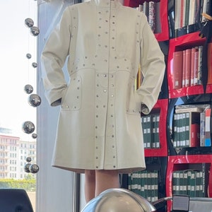 May include: A white leather coat with silver studs. The coat has a collar and long sleeves. The coat is on a mannequin in a room with a bookshelf in the background.