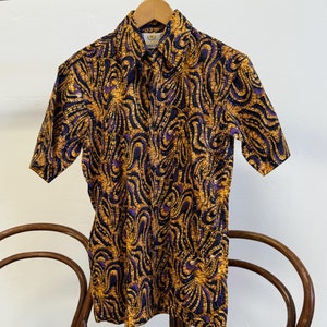 May include: A short-sleeved button-down shirt with a colourful abstract pattern in shades of yellow, purple, and black. The shirt is hanging on a wooden hanger.