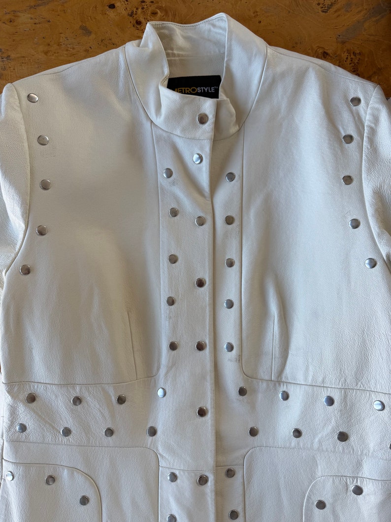 May include: A white leather jacket with silver studs. The jacket has a mandarin collar and a button closure. The label on the jacket reads "Metrostyle".