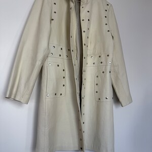 May include: A long, off-white leather jacket with silver studs. The jacket has a stand-up collar, a zipper closure, and two pockets. The jacket is on a hanger.