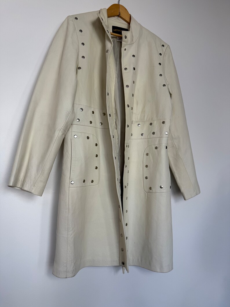 May include: A long, off-white leather jacket with silver studs. The jacket has a stand-up collar, a zipper closure, and two pockets. The jacket is on a hanger.