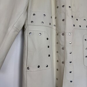 May include: A white leather coat with silver studs. The coat has a long, fitted silhouette and a stand-up collar. The studs are arranged in a geometric pattern on the front and back of the coat.