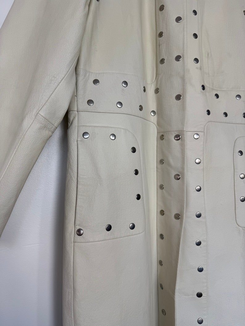 May include: A white leather coat with silver studs. The coat has a long, fitted silhouette and a stand-up collar. The studs are arranged in a geometric pattern on the front and back of the coat.