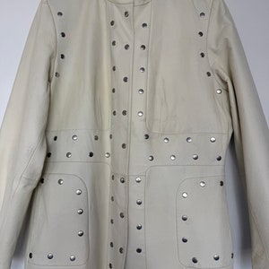 May include: A white leather jacket with silver studs. The jacket has a high collar and a long, fitted silhouette.