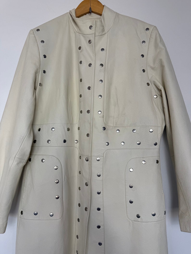 May include: A white leather jacket with silver studs. The jacket has a high collar and a long, fitted silhouette.