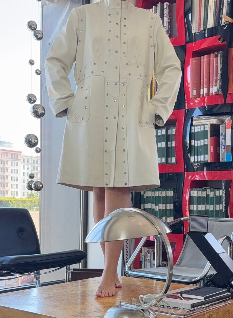 May include: A white leather coat with silver studs. The coat has a collar and long sleeves. The coat is on a mannequin in a room with a bookshelf in the background.