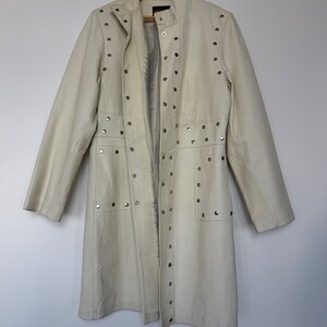 May include: A white leather jacket with silver studs. The jacket has a long, fitted silhouette and a stand-up collar.