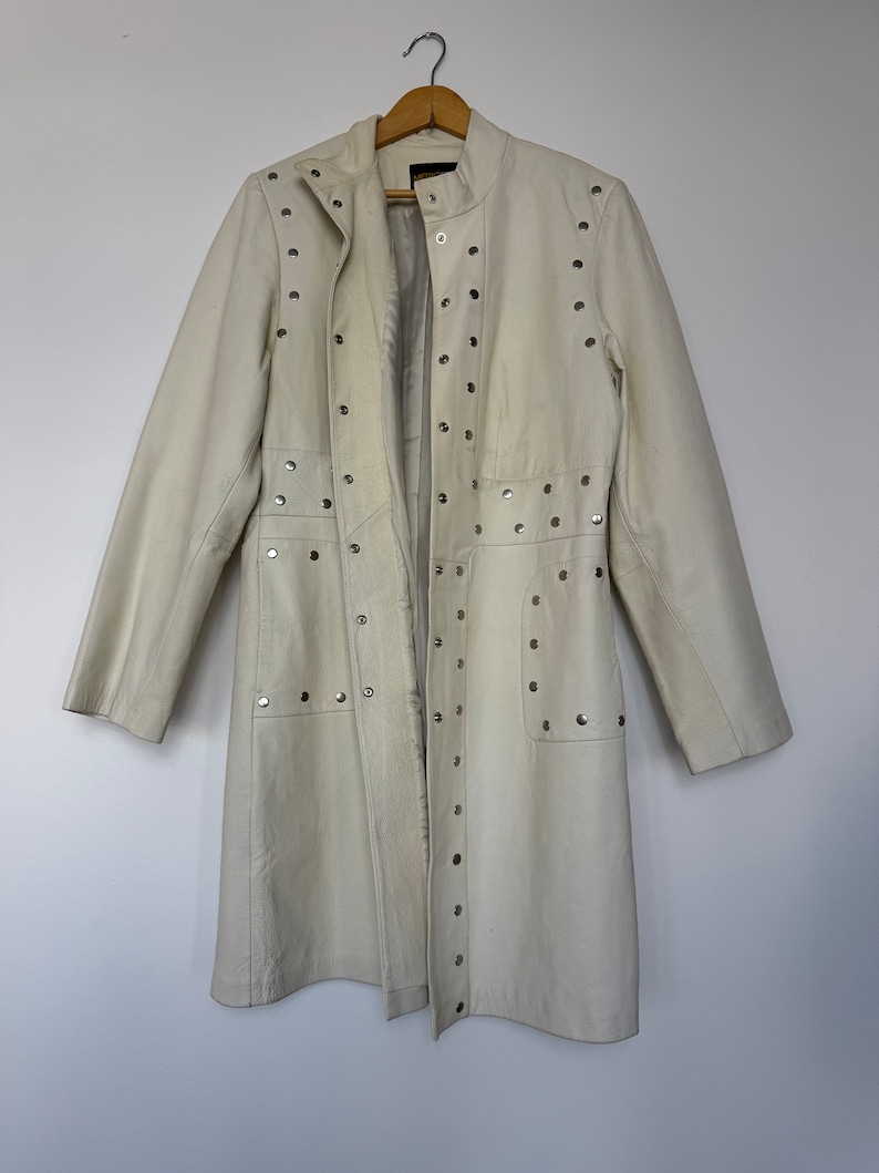 May include: A white leather jacket with silver studs. The jacket has a long, fitted silhouette and a stand-up collar.