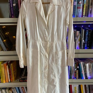 May include: A vintage, off-white dress with long sleeves, a collared neckline, and a button-down front. The dress features two front pockets and is hanging on a wooden hanger. The dress is displayed in front of a bookshelf.