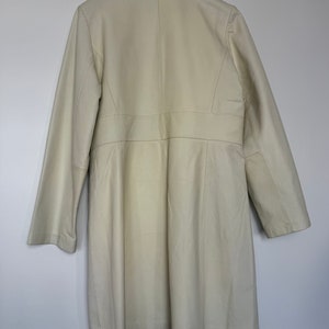 May include: A long, off-white coat with a collar and long sleeves. The coat is hanging on a wooden hanger.