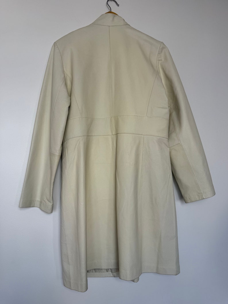 May include: A long, off-white coat with a collar and long sleeves. The coat is hanging on a wooden hanger.