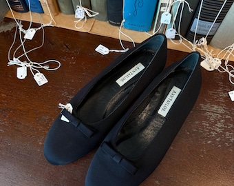 May include: A pair of black Ann Taylor flats with small bow details. The shoes are displayed on a wooden surface, with a backdrop of vintage books. The Ann Taylor label is visible inside each shoe.