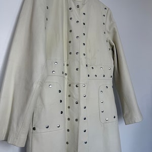 May include: A long, off-white leather coat with silver studs. The coat has a stand-up collar and long sleeves.