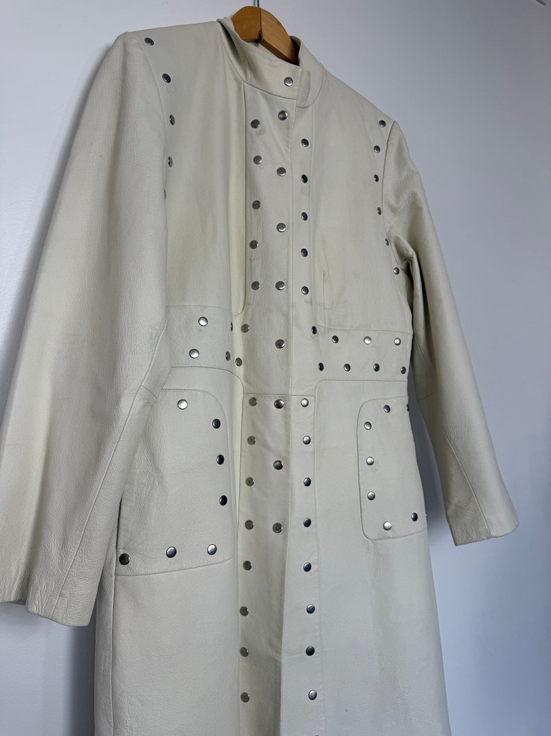 May include: A long, off-white leather coat with silver studs. The coat has a stand-up collar and long sleeves.