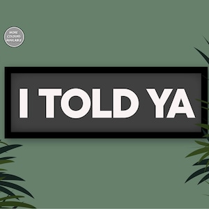 May include: A black framed sign with white text that reads "I TOLD YA" against a green background with green leafy plants on either side.