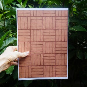 May include: A rectangular sheet of paper featuring a repeating pattern of brown, rectangular blocks resembling a wood grain design. The paper is held against a backdrop of green foliage.