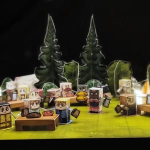 Puede incluir: Un diorama con figuras de papel alrededor de mesas, con árboles y tiendas de campaña al fondo. Las figuras tienen diversas expresiones y parecen estar acampando. La escena se desarrolla sobre una superficie verde, con un fondo oscuro.