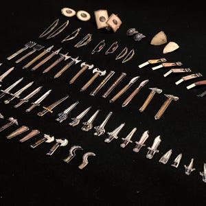 May include: A collection of miniature fantasy weapons, including swords, axes, hammers, and bows, arranged on a black surface. The weapons vary in size and design, with silver and brown tones. Some have shields and other accessories.