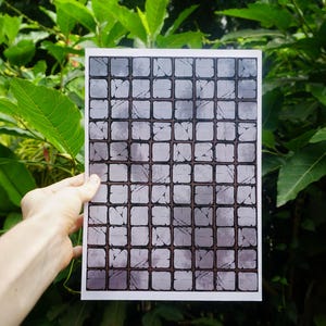 Op de afbeelding: Een rechthoekige print met een raster van grijze, vierkante tegels met een textuur die op steen lijkt. De tegels hebben donkere omtrekken en een enigszins verweerde uitstraling. De print wordt tegen een achtergrond van groen gebladerte gehouden.