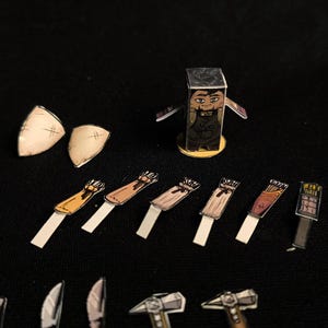 May include: A collection of paper cutouts of fantasy game pieces, including a character, shields, quivers, and weapons. The character is a rectangular figure with a face and a dark outfit. The pieces are arranged on a black surface.