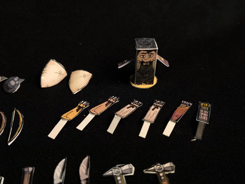 May include: A collection of paper cutouts of fantasy game pieces, including a character, shields, quivers, and weapons. The character is a rectangular figure with a face and a dark outfit. The pieces are arranged on a black surface.