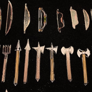 May include: A collection of miniature toy weapons, including bows, arrows, spears, and axes, arranged on a black surface. The bows vary in design, with some featuring a level. The axes have double-sided blades.