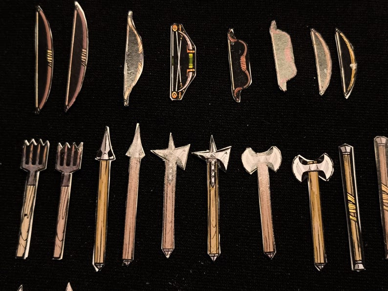 May include: A collection of miniature toy weapons, including bows, arrows, spears, and axes, arranged on a black surface. The bows vary in design, with some featuring a level. The axes have double-sided blades.