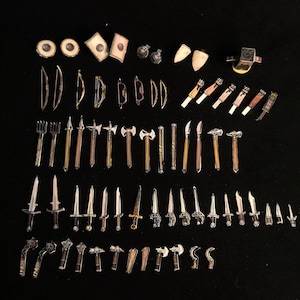 May include: A collection of miniature fantasy weapons and accessories, including swords, axes, bows, and shields, arranged on a black surface. The items are made of metal and wood, with varying colors and designs. The assortment includes small hammers and knives.