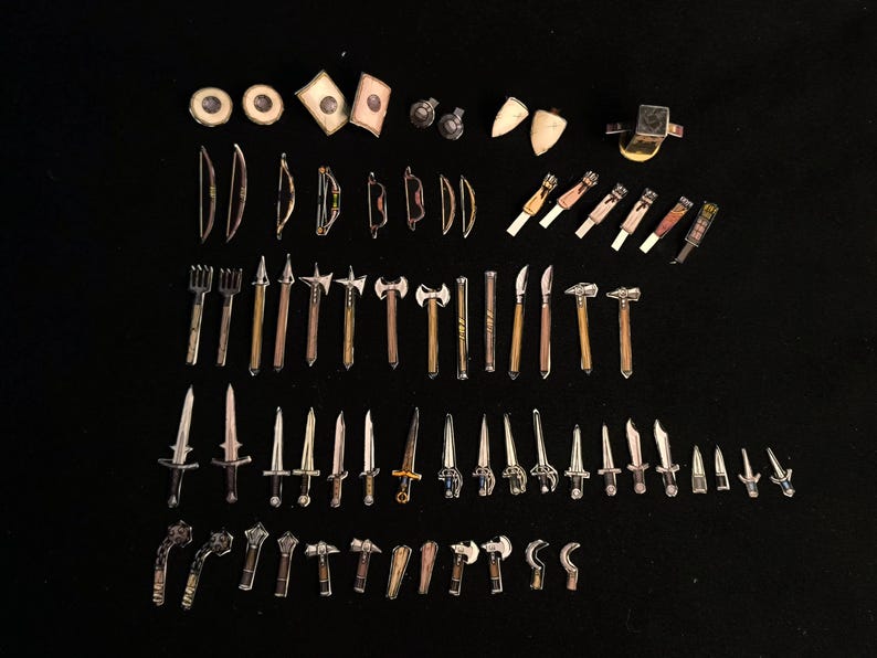 May include: A collection of miniature fantasy weapons and accessories, including swords, axes, bows, and shields, arranged on a black surface. The items are made of metal and wood, with varying colors and designs. The assortment includes small hammers and knives.