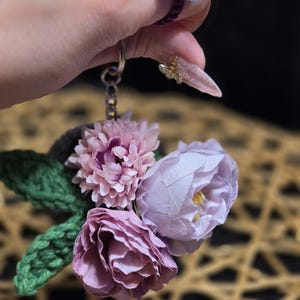 May include: A keychain featuring a cluster of three artificial fabric flowers in shades of dusty pink and light purple, accented with two crocheted green leaves. The floral arrangement is attached to a silver-toned metal chain and ring.