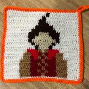 May include: A crocheted square with an orange border featuring a pixelated image of a person with brown hair wearing a red and brown outfit.